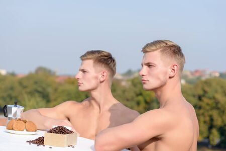 young handsome twins with sexy muscular athletic strong body has bare torso and strong belly having breakfast on roof with serious face on blue sky backgroundの写真素材