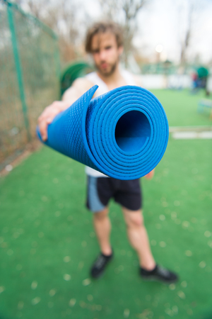 mat. yoga or fitness mat blue color in hands of man with muscular body and beard exercise in outdoor gym or arena in sportswear, energetic handsome guy or sexy bearded macho workoutの写真素材