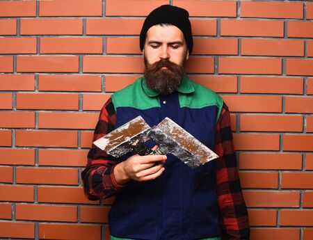bearded worker man, long beard, brutal caucasian hipster with moustache in uniform holding spatulas with serious face on brick wall studio backgroundの写真素材