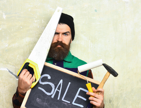 bearded man, long beard, brutal caucasian hipster with moustache holding various building tools: saw, hammer, roller paint and board with inscription sale with serious face on studio wall backgroundの写真素材