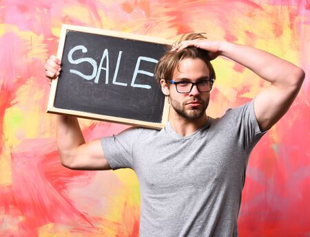 Bearded man, short beard. Caucasian sexy serious macho man with moustache in glasses wearing gray shirt holding black board with sale inscription on colorful studio backgroundの写真素材