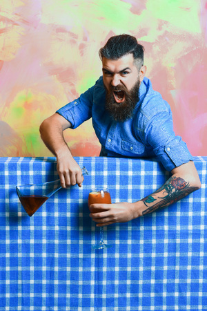 Bearded man, long beard. Brutal caucasian shouting hipster with moustache in denim shirt holding flask or bottle doing tropical alcohol cocktail on blue checkered plaid on colorful texture backgroundの写真素材
