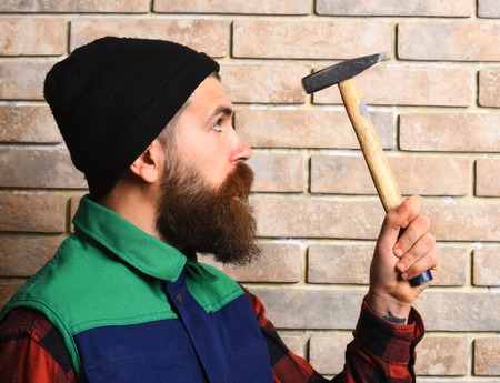 bearded builder man, long beard, brutal caucasian hipster with moustache in black cap holding hammer with serious face on brick wall studio backgroundの写真素材