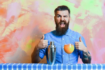 Bearded man, long beard. Brutal caucasian happy hipster with moustache in denim shirt doing tropical alcohol fresh cocktail with shaker on blue checkered plaid on colorful texture backgroundの写真素材