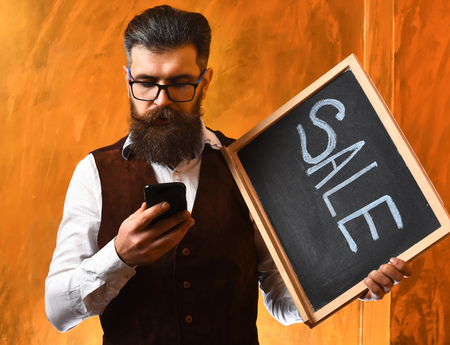 Bearded man, long beard. Brutal caucasian unshaven serious hipster with glasses and moustache surfing internet on cell phone, holding sale inscription on blackboard on brown studio backgroundの写真素材