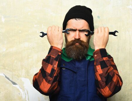 Bearded man, long beard. Brutal caucasian mechanic, unshaven hipster in black hat, checkered shirt and uniform holding metallic wrenches on texture studio backgroundの写真素材