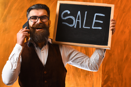 Bearded man, long beard. Brutal caucasian unshaven amused hipster with glasses and moustache talking by cell phone, holding sale inscription on blackboard on brown studio backgroundの写真素材