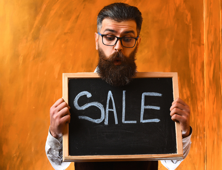 Bearded man, long beard. Brutal caucasian unshaven surprised hipster with glasses and moustache holding sale inscription on blackboard on brown studio backgroundの写真素材