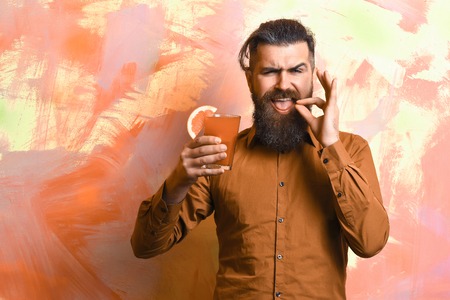 Bearded man, long beard. Brutal caucasian satisfied hipster with moustache in brown shirt holding tropical alcoholic fresh cocktail with orange piece on colorful texture backgroundの写真素材