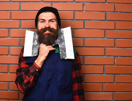 bearded worker man, long beard, brutal caucasian hipster with moustache in uniform holding spatulas with happy smiling face on brick wall studio backgroundの写真素材
