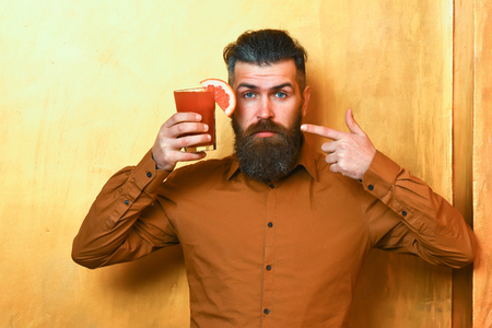 Bearded man, long beard. Brutal caucasian serious hipster with moustache in brown shirt holding tropical alcoholic fresh cocktail with orange piece on golden texture backgroundの写真素材