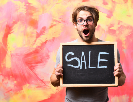 Bearded man, short beard. Caucasian sexy surprised macho man with moustache in glasses wearing gray shirt holding black board with sale inscription on colorful studio backgroundの写真素材
