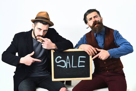 Two bearded mens, short and long beard. Caucasian stylish hipsters with moustache holding sale inscription on blackboard and smoking cigars, isolated on white backgroundの写真素材