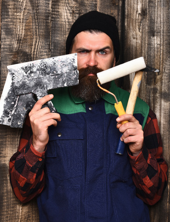 bearded builder man, long beard, brutal caucasian hipster with moustache in uniform holding various building tools: hammer, roller paint, spatulas with serious face on brown wooden studio backgroundの写真素材