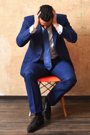 tired young fashion business man in classy suit and tie sitting on chair on beige background, stress and overtimeの写真素材
