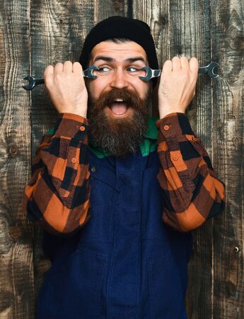 Bearded man, long beard. Brutal caucasian mechanic, smiling happy unshaven hipster in black hat, checkered shirt and uniform holding metallic wrenches on brown vintage wooden studio backgroundの写真素材