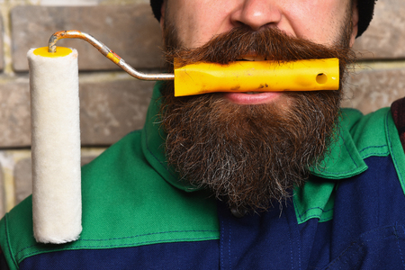 bearded painter man, long beard, brutal caucasian hipster with moustache in black cap holding in his mouth roller paint with serious face on brick wall studio background, closeupの写真素材