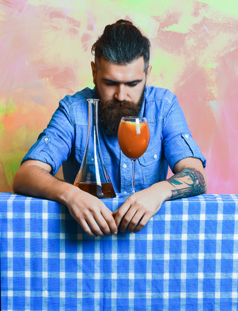 Bearded man, long beard. Brutal caucasian tattooed hipster with moustache in denim shirt with tropical alcohol fresh cocktail and flask on blue checkered plaid on colorful texture backgroundの写真素材