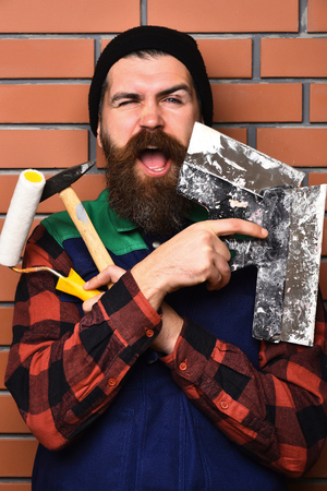 bearded builder man, long beard, brutal caucasian hipster with moustache in uniform holding various building tools: hammer, roller paint and spatulas with happy, face on brick wall studio backgroundの写真素材