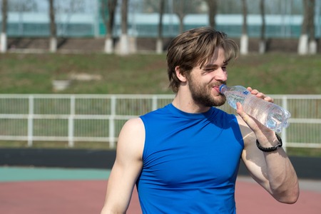 handsome man or guy with muscular body holds plastic refreshing water bottle sunny outdoor in blue sportswear at stadium. fitness and healthy lifestyle conceptの写真素材