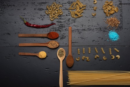 Pasta and spices. Cooking composition made of differents sorts of pasta with spices in wooden spoons and chili pepper on dark grey and rough surface, top viewの写真素材