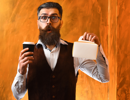 Bearded man, long beard. Brutal caucasian unshaven surprised hipster with glasses and moustache holding black plastic coffee cup or mug and meal box on brown studio backgroundの写真素材