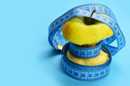 Tangled bitten apple. Blue flexible ruler measures green apple tying it around on blue background, close up and copy spaceの写真素材