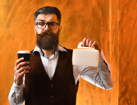 Bearded man, long beard. Brutal caucasian unshaven serious hipster with glasses and moustache holding black plastic coffee cup or mug and meal box on brown studio backgroundの写真素材