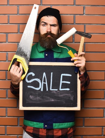 bearded man, long beard, brutal caucasian hipster with moustache holding various building tools: saw, hammer, roller paint and board with inscription sale with serious face on brick wall backgroundの写真素材