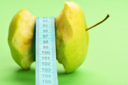 Concept of organic food, healthy nutrition and vegetarian lifestyle: bitten apple lying on side tied around with cyan measuring tape on light green background with copy space, selective focusの写真素材