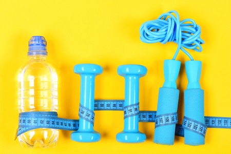 Gym and sports concept with bottle of water, dumbbells and skipping rope wrapped with blue measuring tape isolated on yellow background, top viewの写真素材