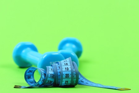 Measuring tape in blue colour and light blue dumbbells behind, isolated on green background, close up with selective focus and copy space. Sports and fit shape conceptの写真素材