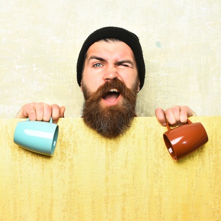 Bearded man, long beard. Brutal caucasian funny unshaven hipster in black hat with yellow paper sheet holding colorful cups on texture studio background, copy spaceの写真素材