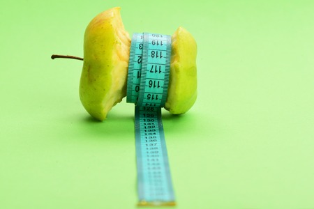 Fat loss concept. bitten apple lying on side belted with cyan measuring tape on light green background with copy spaceの写真素材