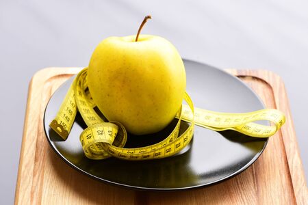 Apple, big ripe and yellow, standing on black ceramic plate with yellow measuring tape placed on wooden cutting board on grey background, selective focus. Diet conceptの写真素材