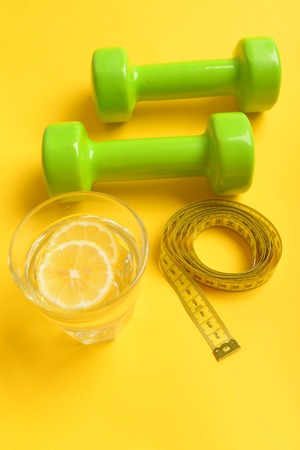 Health and sports concept with green dumbbells, yellow measuring tape and lemon water in glass isolated on yellow background, top viewの写真素材