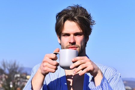 Bearded man, short beard. Caucasian macho with moustache and ruffled hair in blue striped pajamas holding cup or mug in roof on blurred city background, morning breakfast conceptの写真素材