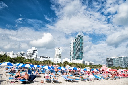 Miami, USA - January 10, 2016: Miami beach or south beach. people sunbathing on deck chairs under blue umbrellas on beach with high buildings on cloudy blue sky. Summer vacation. Lounge and leisureのeditorial素材