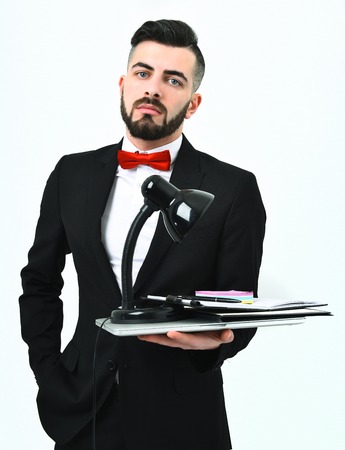 Bearded office manager in black suit and red bow holding laptop with lamp and stationery on it, isolated on white background. Confident and successful business and office work conceptの写真素材