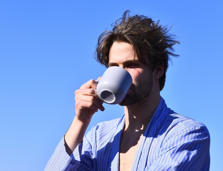 Bearded man, short beard. Caucasian macho with moustache and ruffled hair in blue striped pajamas holding cup or mug in roof on blue sunny sky background, morning coffee conceptの写真素材