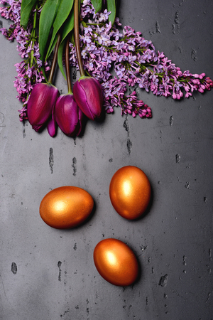 Spring bouquet of tulips and lilac near golden eggs for Easter on rough grey stone background, top view. Easter decorations and flowersの写真素材