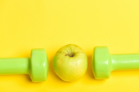 Dumbbells in juicy green colour and ripe greenish yellow apple between them isolated on yellow background, copy space. Concept of healthy nutrition and sportive shapeの写真素材