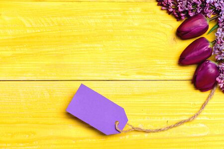 Label card in purple color tied to tulips and lilac flowers on yellow wooden background, copy space and top view. Gift for womenの写真素材