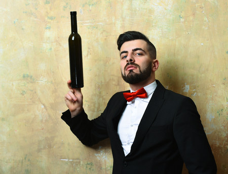 Confident man drinks. Manager holds bottle of alcoholic beverage with his fingers and looks proudly, dirty beige vintage background. Leisure time after workの写真素材