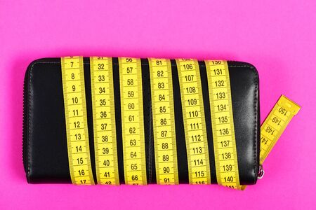 Female purse in black color wrapped with yellow flexible ruler, isolated on pink background, top view. Concept of money savingの写真素材