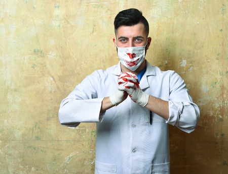 Doctor with smiling eyes and stylish haircut surgical mask and white gloves stained with bloody hands on beige backgroundの写真素材