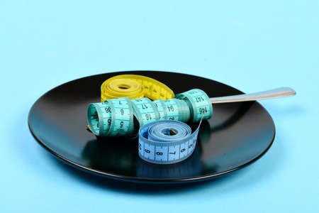 Fork wrapped with cyan measuring tape, rolls of yellow and blue flexible rulers on black plate, isolated on turquoise backgroundの写真素材