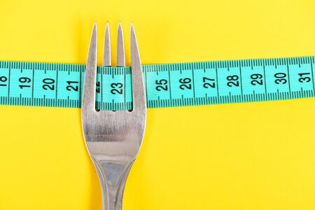 Fork in silver color with four tines holding cyan blue measuring tape on bright yellow background,...