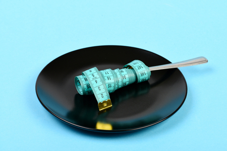 Plate in black color and fork wrapped around with measuring tape on it, isolated on light blue colorの写真素材