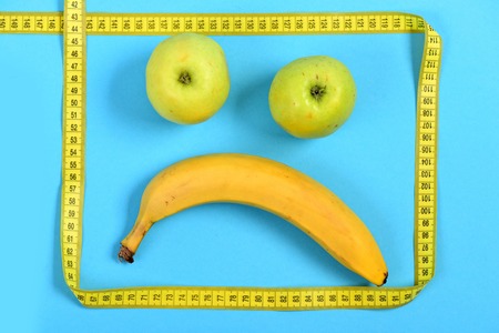Face made of fruit with sad expression: banana and apples making mouth and eyes framed with yellow flexible ruler, isolated on cyan blue background. Concept of food art and minimalismの写真素材